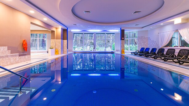 Une grande piscine intérieure à l’eau d’un bleu limpide, entourée de chaises longues et offrant une vue sur les arbres à travers de grandes fenêtres.