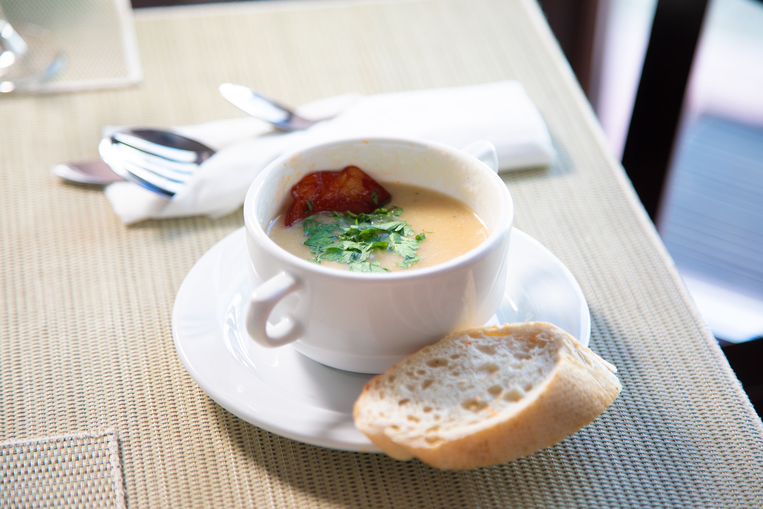 A white cup of soup with a garnish on a white saucer, accompanied by a slice of bread on a woven placemat.