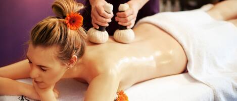 A massage therapy session in progress with a client lying face down on a massage table, a therapist applying pressure with massage tools, and a flower on the table.