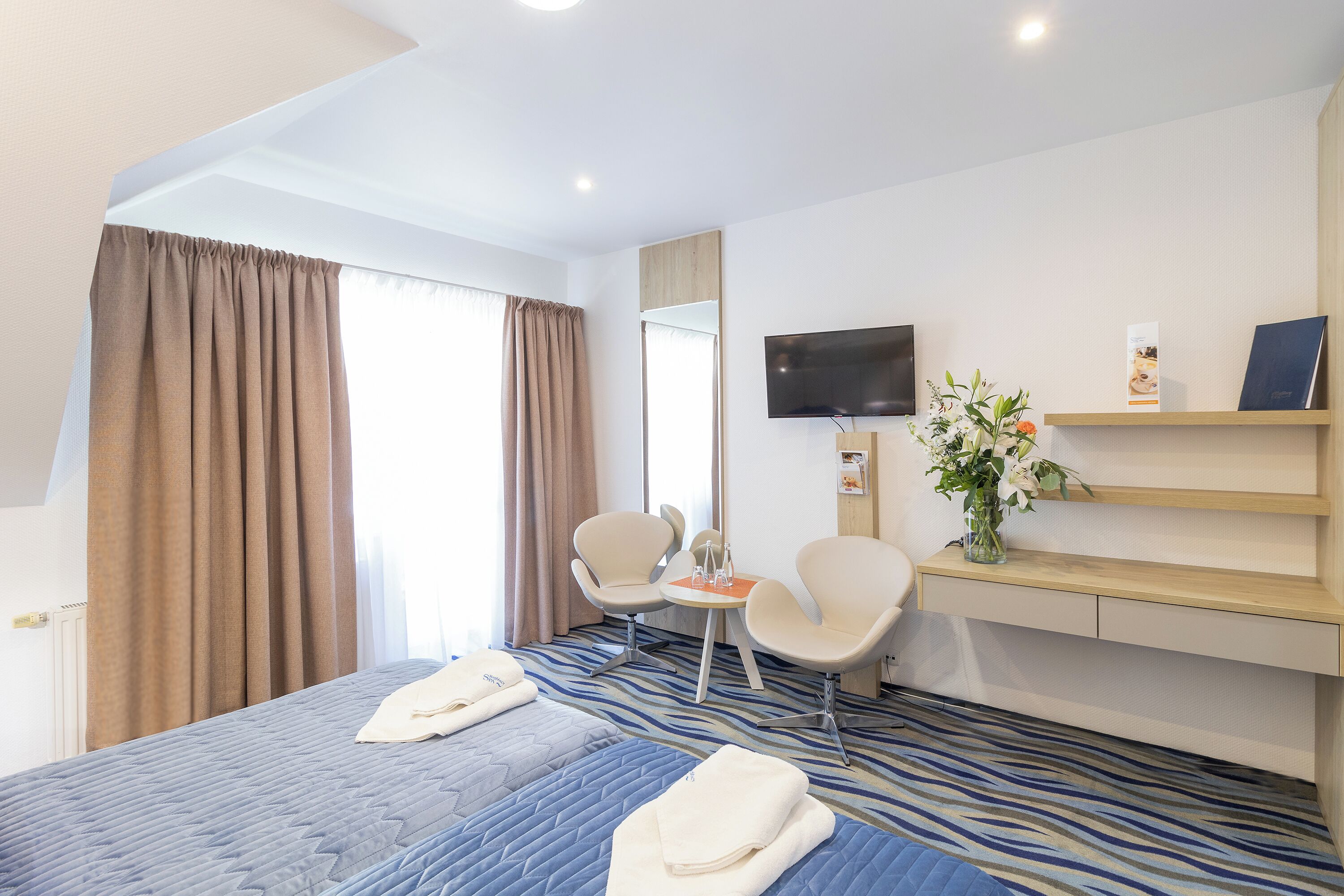 A hotel room with a bed, two chairs, a small table, a TV, and a shelf with a vase of flowers.