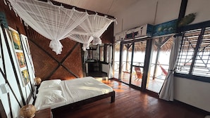 A room with a bed, wooden flooring, a hanging canopy, artwork on the walls, and a view through large windows. - Waterfront villa with live-in staff! (NOSY BE)