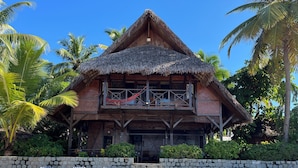 A wooden building with a thatched roof, a balcony with a hammock, and a stone wall with lounge chairs in front. - Waterfront villa with live-in staff! (NOSY BE)