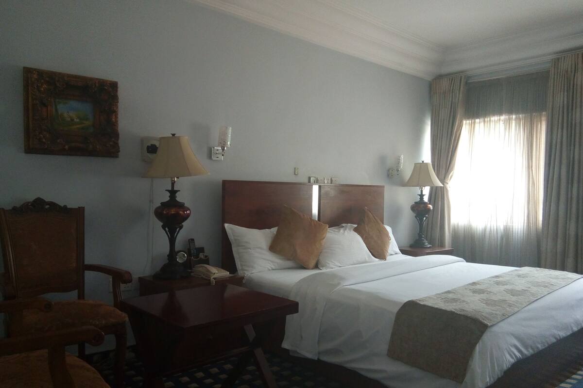 Une chambre d’hôtel avec un lit, deux tables de chevet, une chaise, une lampe, un tableau au mur et une fenêtre avec des rideaux.