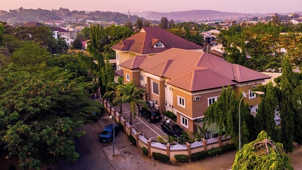 Une grande maison à deux étages, avec un toit recouvert de tuiles rouges, entourée d’une végétation luxuriante et dotée d’une allée pavée.