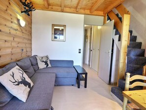 A modern interior with a grey sectional sofa, a wooden headboard, a small table, and a staircase with a wooden chair.
