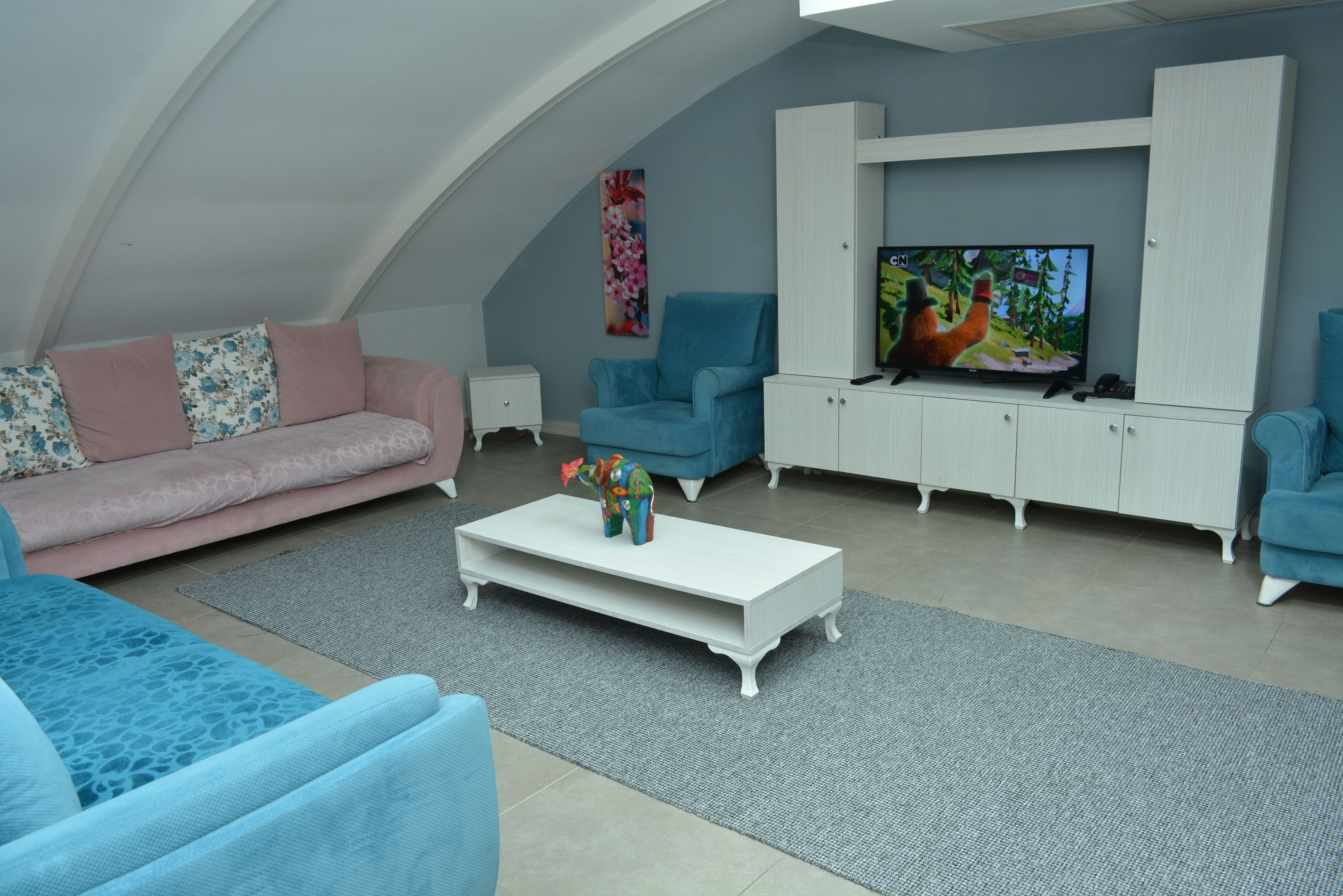 A modern living room with a high ceiling, a television on a white cabinet, a pink sofa, a blue armchair, and a coffee table with decorative items.