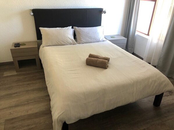 A neatly made bed with a white bedspread and two pillows, a remote control on a bedside table, and a window with curtains.