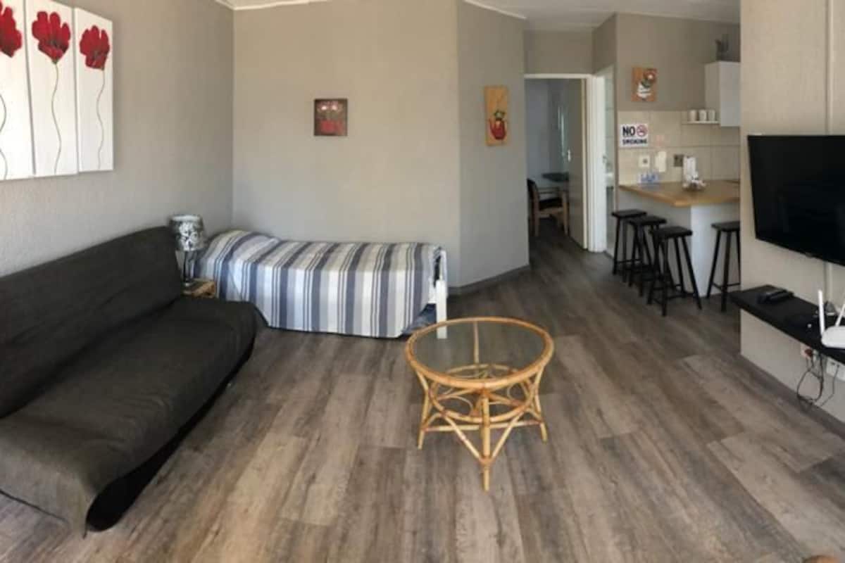 Ein modernes Wohnzimmer mit einer Couch, einem gestreiften Ottomanen, einem runden Korbstuhl, einem an der Wand montierten Fernseher und einem Bett mit einer Zebra-Bettdecke.
