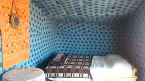 A small, cozy room with a patterned blue and orange wall, a bed with a white and brown blanket, and a small table with a patterned tablecloth. - Locals Under Stars Trips (M'Hamid El Ghizlane)