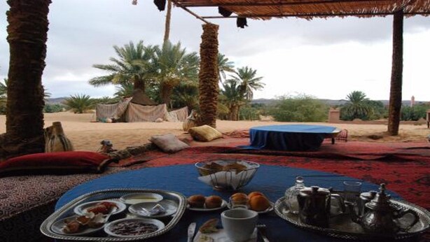 An outdoor dining area with a table set for a meal, surrounded by palm trees and a desert landscape. - Locals Under Stars Trips (M'Hamid El Ghizlane)