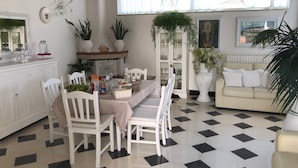 Comedor con una mesa lista para cuatro personas, rodeada de sillas blancas. Hay un sofá blanco y una planta grande en maceta en la esquina.
