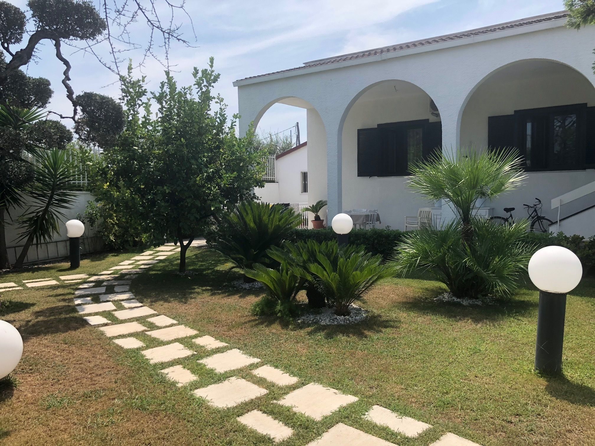 Une cour avec un chemin pavé, un bâtiment blanc orné d’arcades et des plantes en pot.