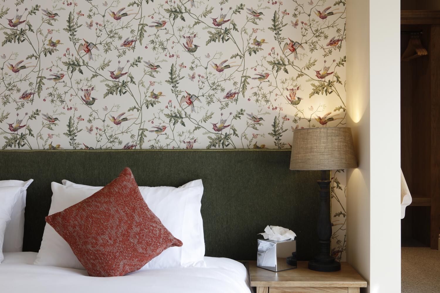 A hotel room with a floral wallpaper, a green headboard, a white bed with a red pillow, a bedside table with a lamp and a tissue box.