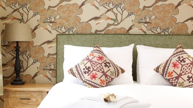 Una cama bien hecha con sábanas blancas y dos almohadones decorativos, una lámpara de noche y una mesita de madera con cajón.