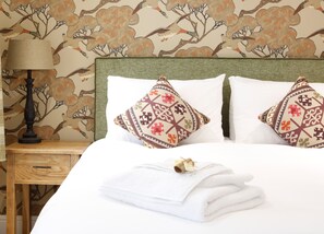 A neatly made bed with white linens and two decorative pillows, a bedside lamp, and a wooden nightstand with a drawer. - Barton's Mill (Basingstoke)