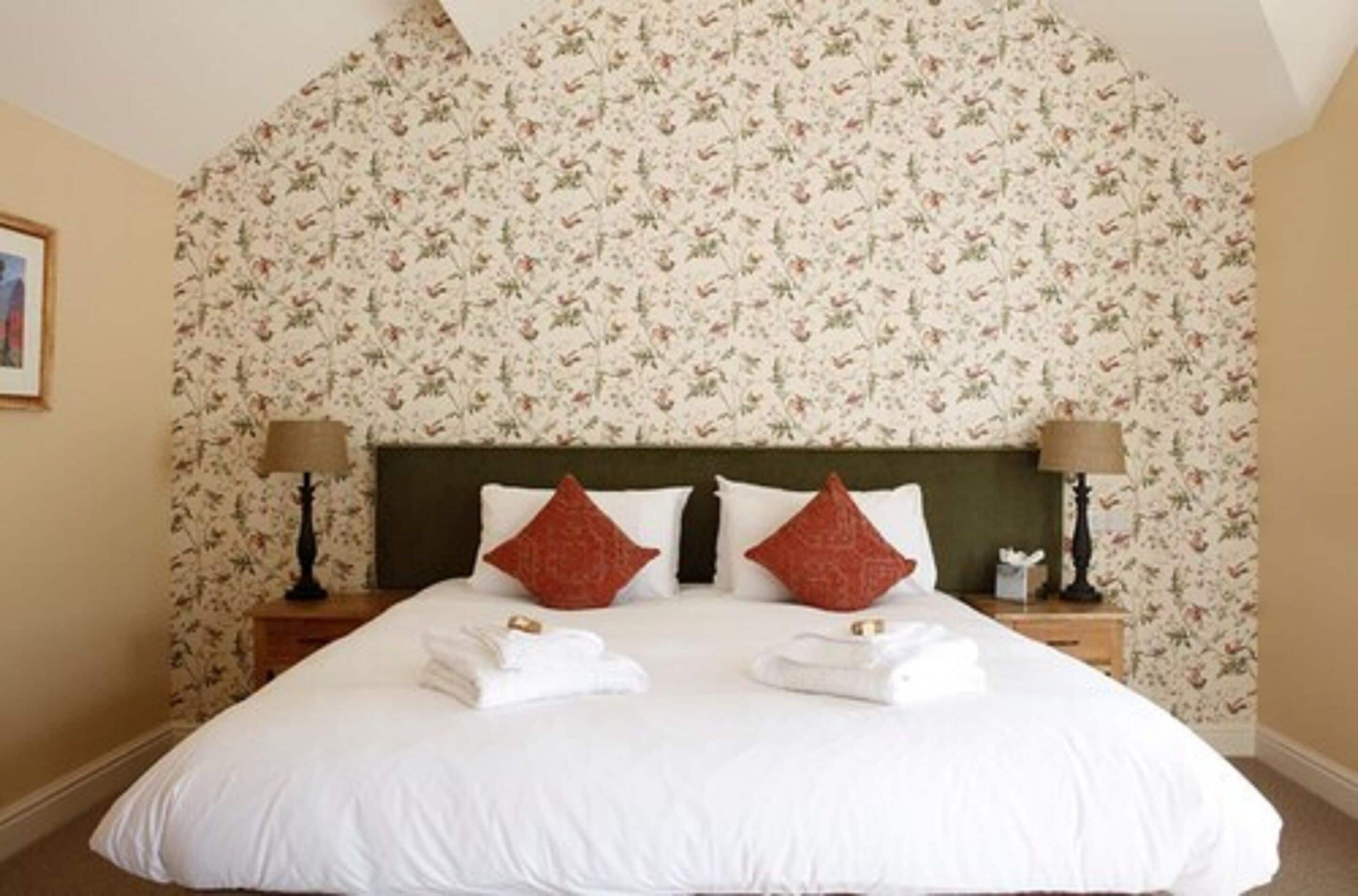 Una cama bien tendida con sábanas blancas, dos almohadas decorativas rojas y un cabecero verde. Hay dos mesitas de noche con lámparas y un pequeño mueble auxiliar con una caja de pañuelos.