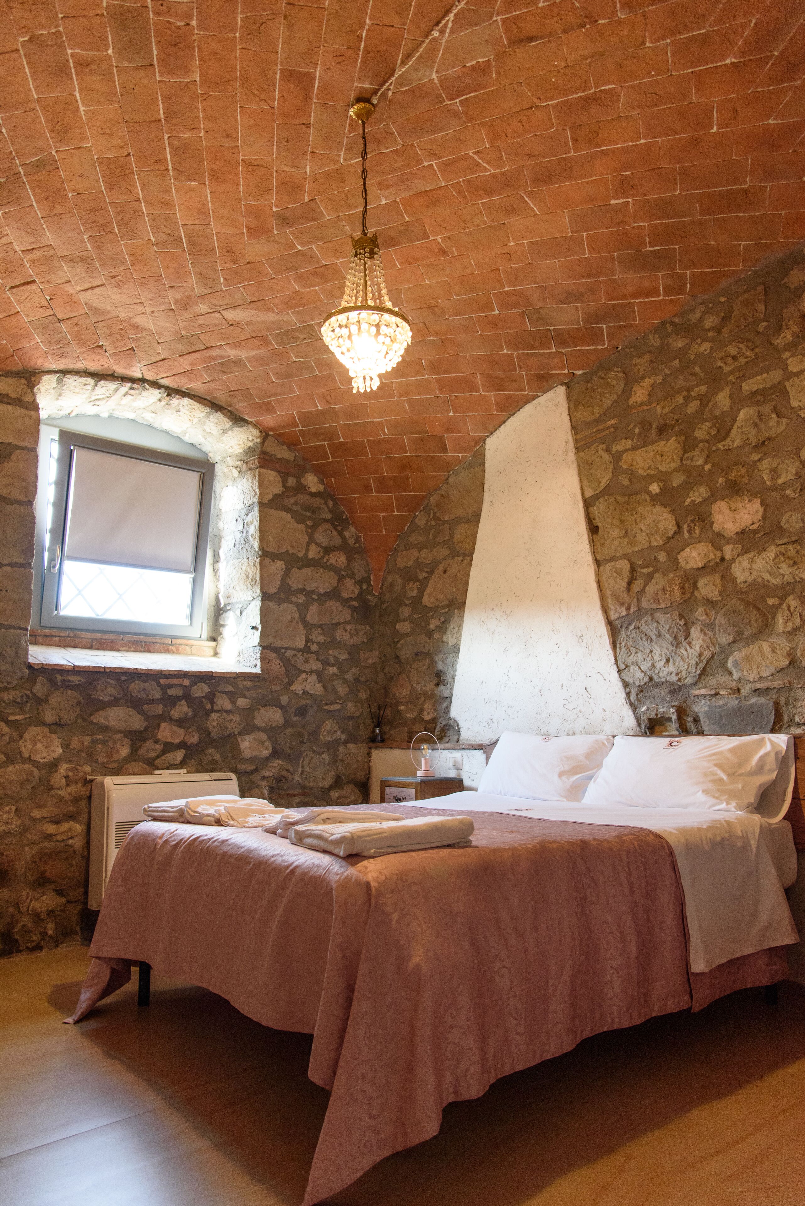Une chambre avec un mur en pierre, une fenêtre, un lustre et un lit recouvert d’une couverture rose.