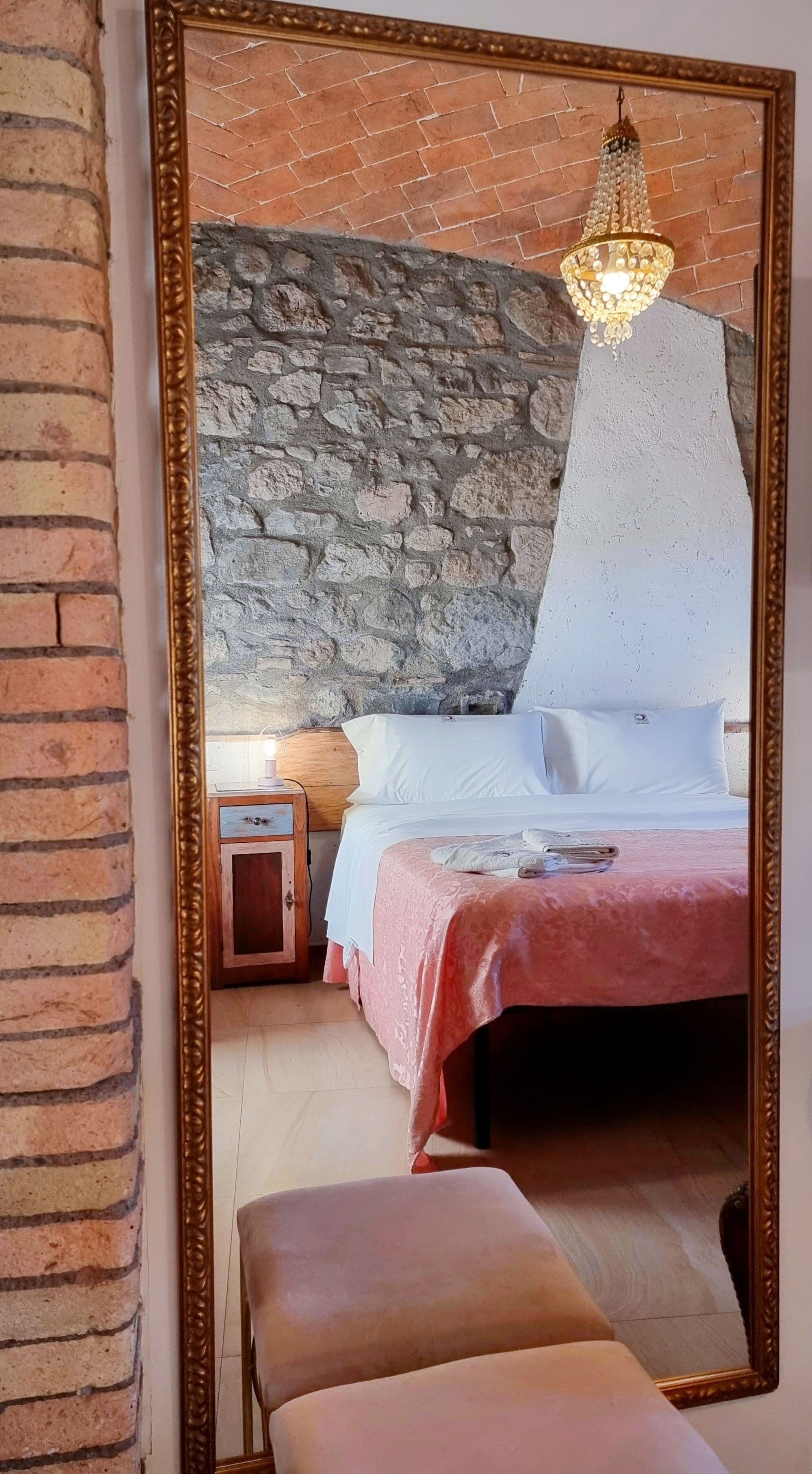 Un dormitorio con pared de piedra, cama con ropa de cama blanca, mesita de noche con lámpara y un espejo grande.