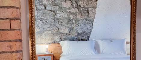 Un dormitorio con pared de piedra, cama con ropa de cama blanca, mesita de noche con lámpara y un espejo grande.