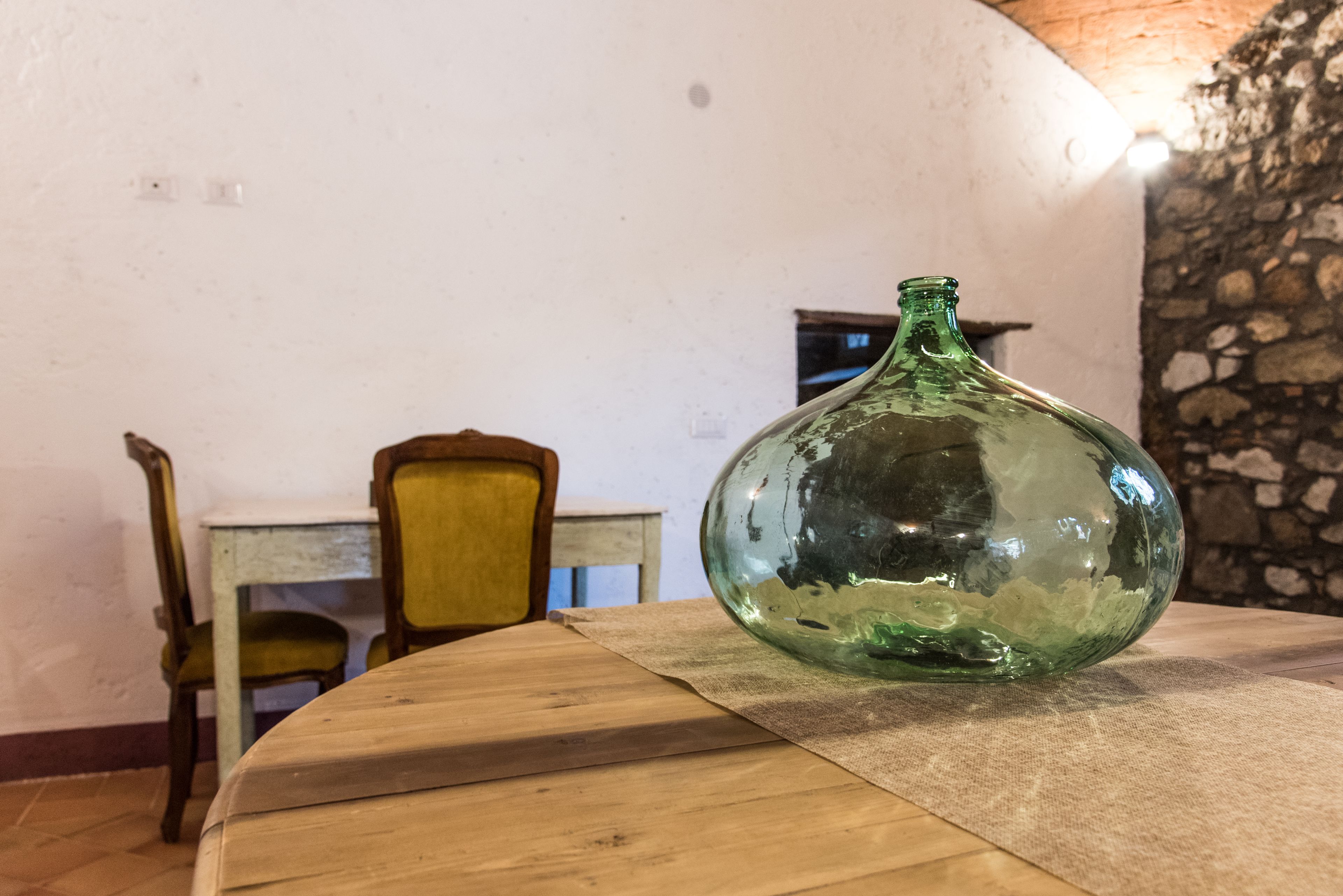 Una botella de vidrio verde sobre una mesa de madera en una habitación con pared de piedra y una silla amarilla.