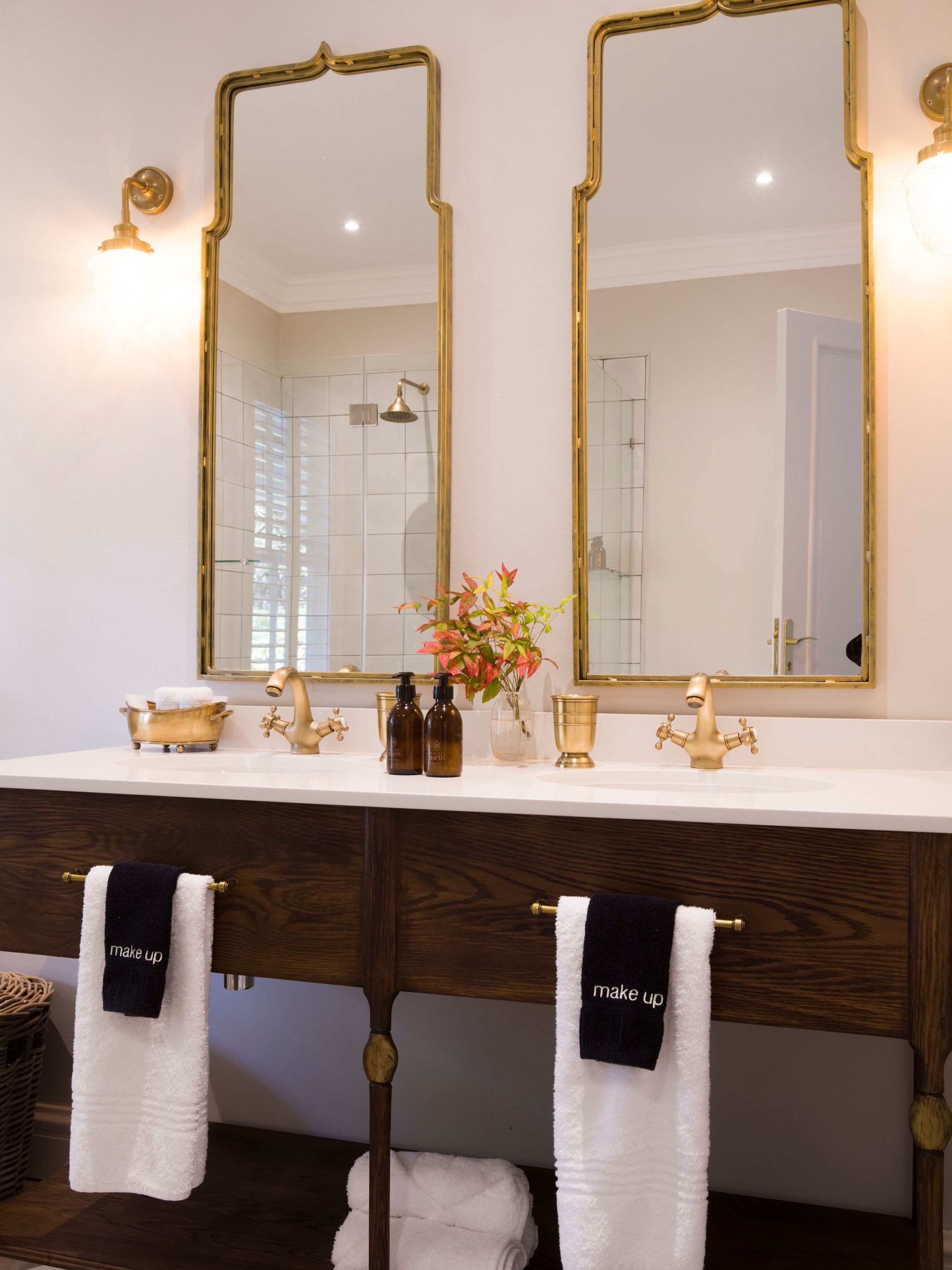 Une salle de bain avec un lavabo blanc, deux robinets dorés et deux miroirs avec des cadres dorés. Des serviettes sont suspendues à un porte-serviettes en bois, et un vase rempli de fleurs est posé sur le lavabo.
