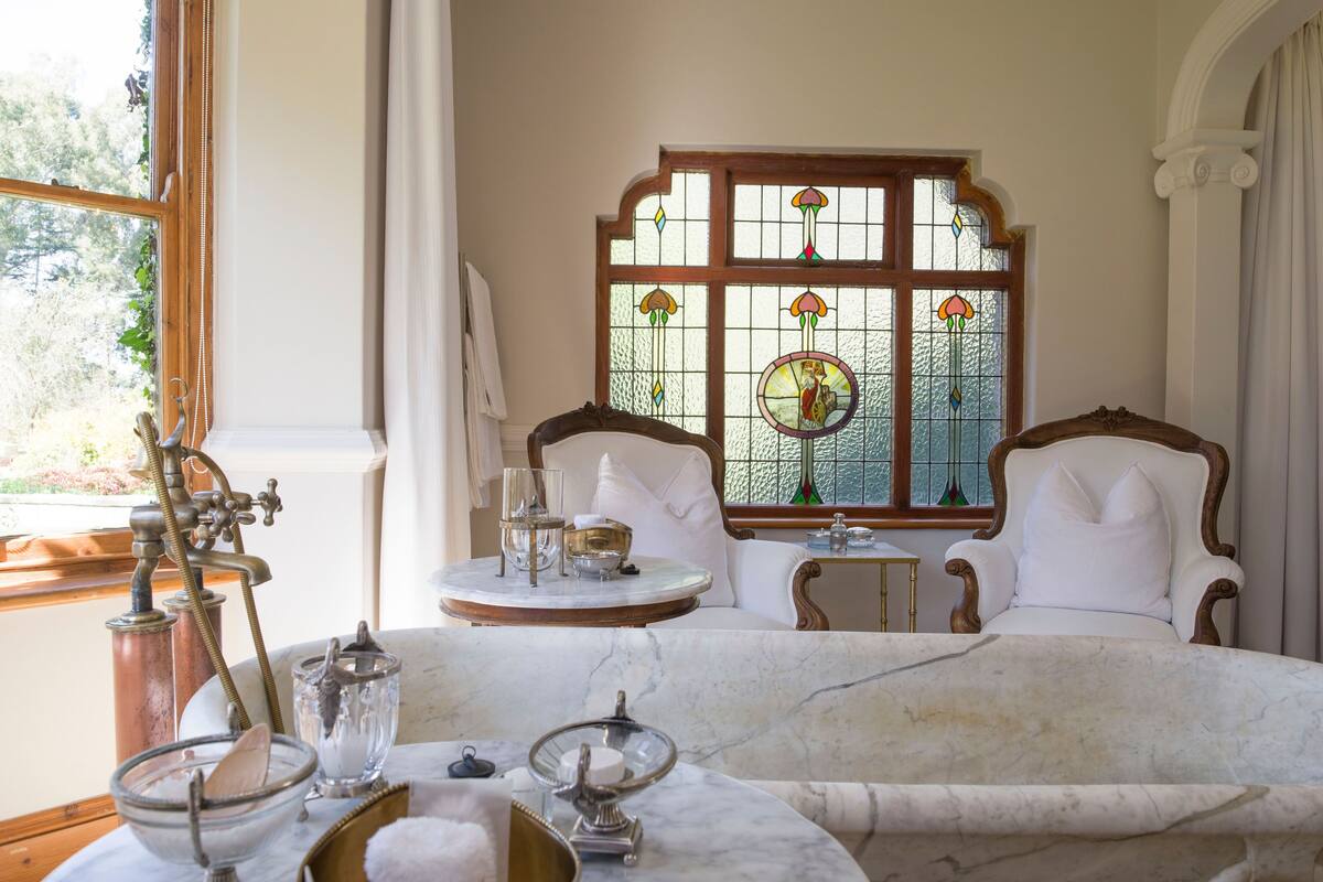 Een luxe badkamer met een ruime badkuip, twee beklede witte fauteuils, een klein tafeltje met toiletartikelen en een glas-in-loodraam.