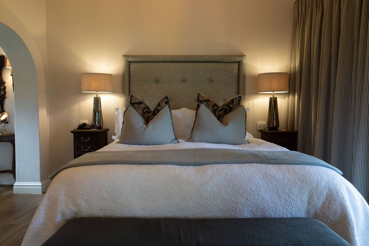 Een netjes opgemaakt bed met een gestoffeerd hoofdbord, twee bedlampen en een groot raam met gordijnen.