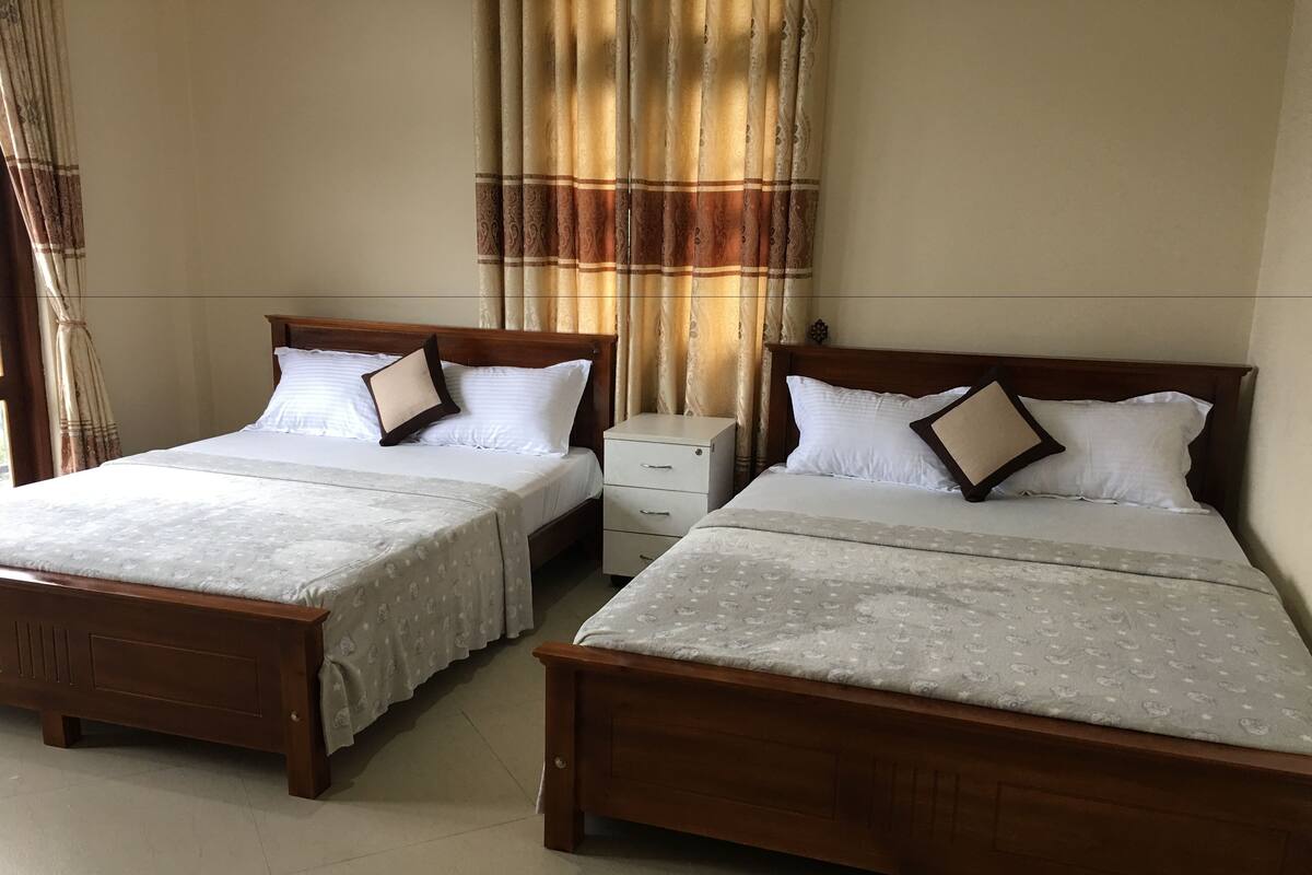 Two wooden beds with white linens and patterned bedspreads, a bedside table, and a window with curtains.