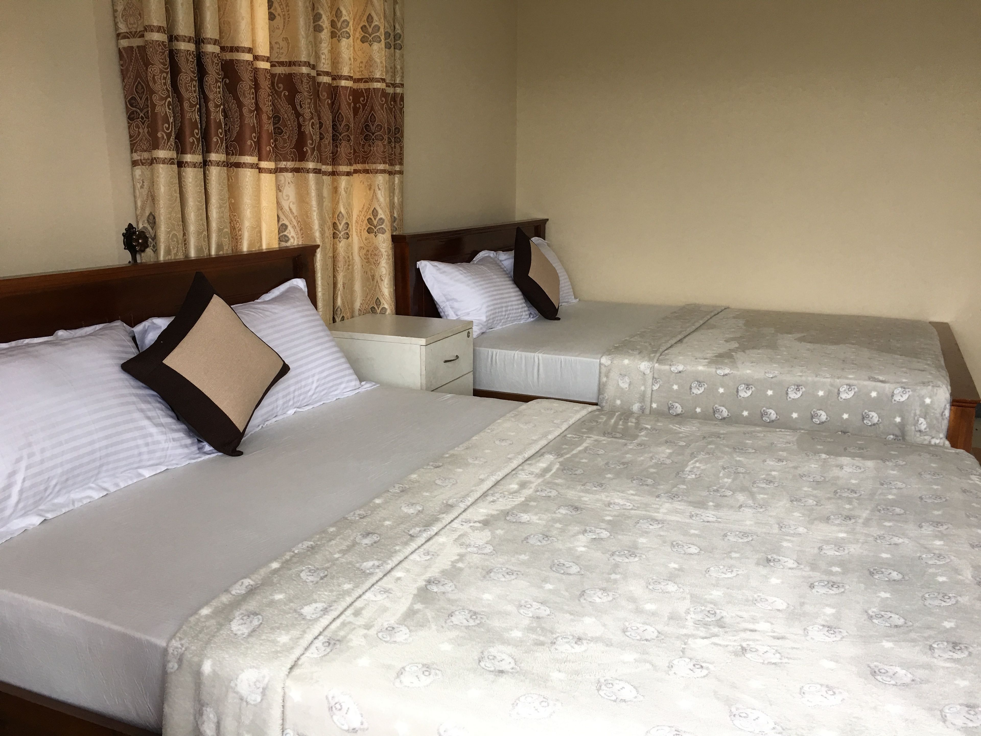 Dos camas individuales con ropa blanca y mantas estampadas, una mesita de noche de madera y una cortina decorativa.