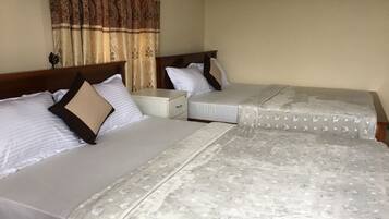 Dos camas individuales con ropa blanca y mantas estampadas, una mesita de noche de madera y una cortina decorativa.