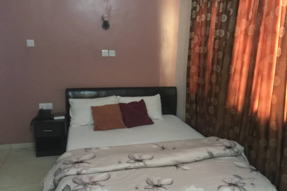 A bedroom with a bed, a nightstand, a wall socket, and a curtain.