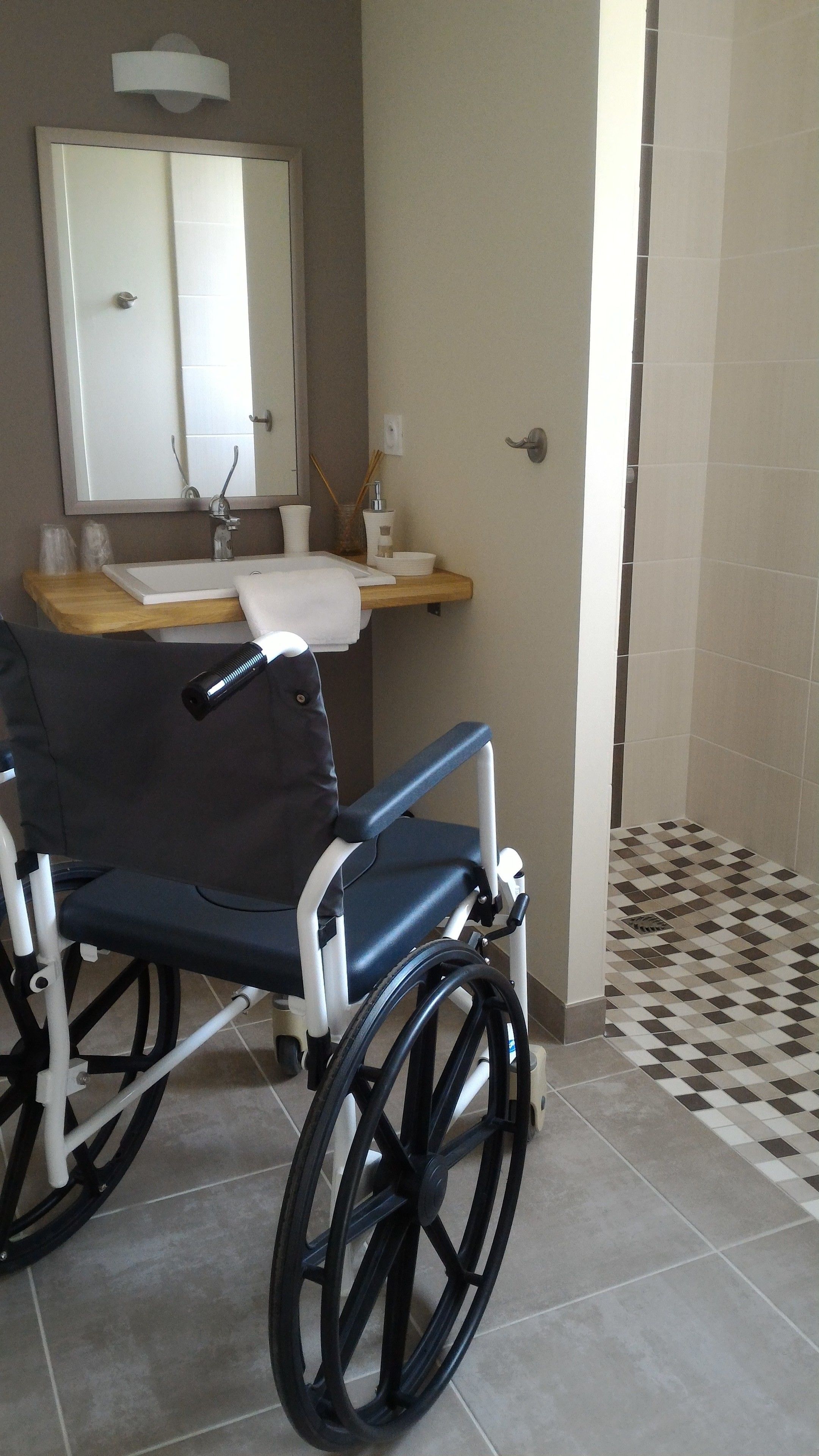 Un fauteuil roulant dans une salle de bain équipée d’un lavabo, d’un miroir et d’un sol carrelé.