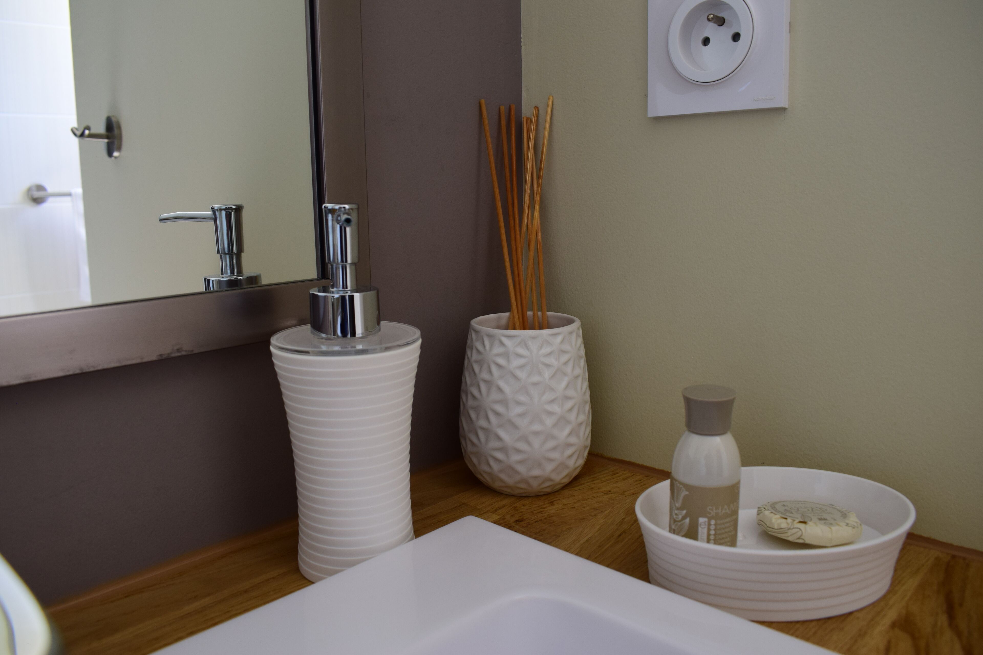 Een badkamer met een wit wastafelmeubel, een spiegel, een witte vaas met rietstelen en een witte kom met een fles en zeep.