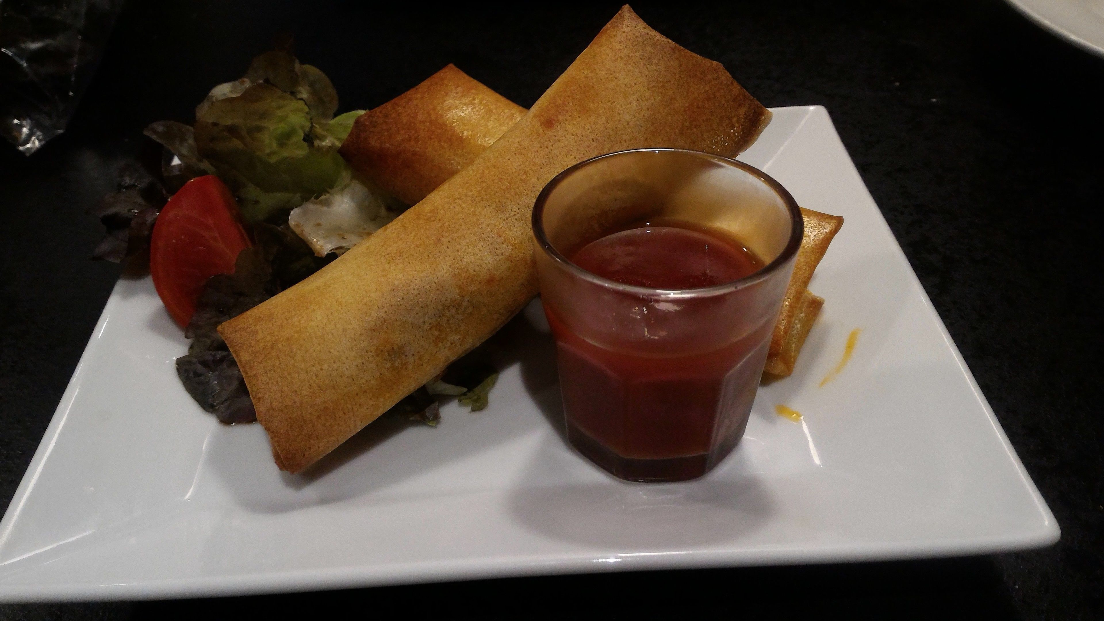 A plate with spring rolls, a glass of ketchup, and a slice of bread.