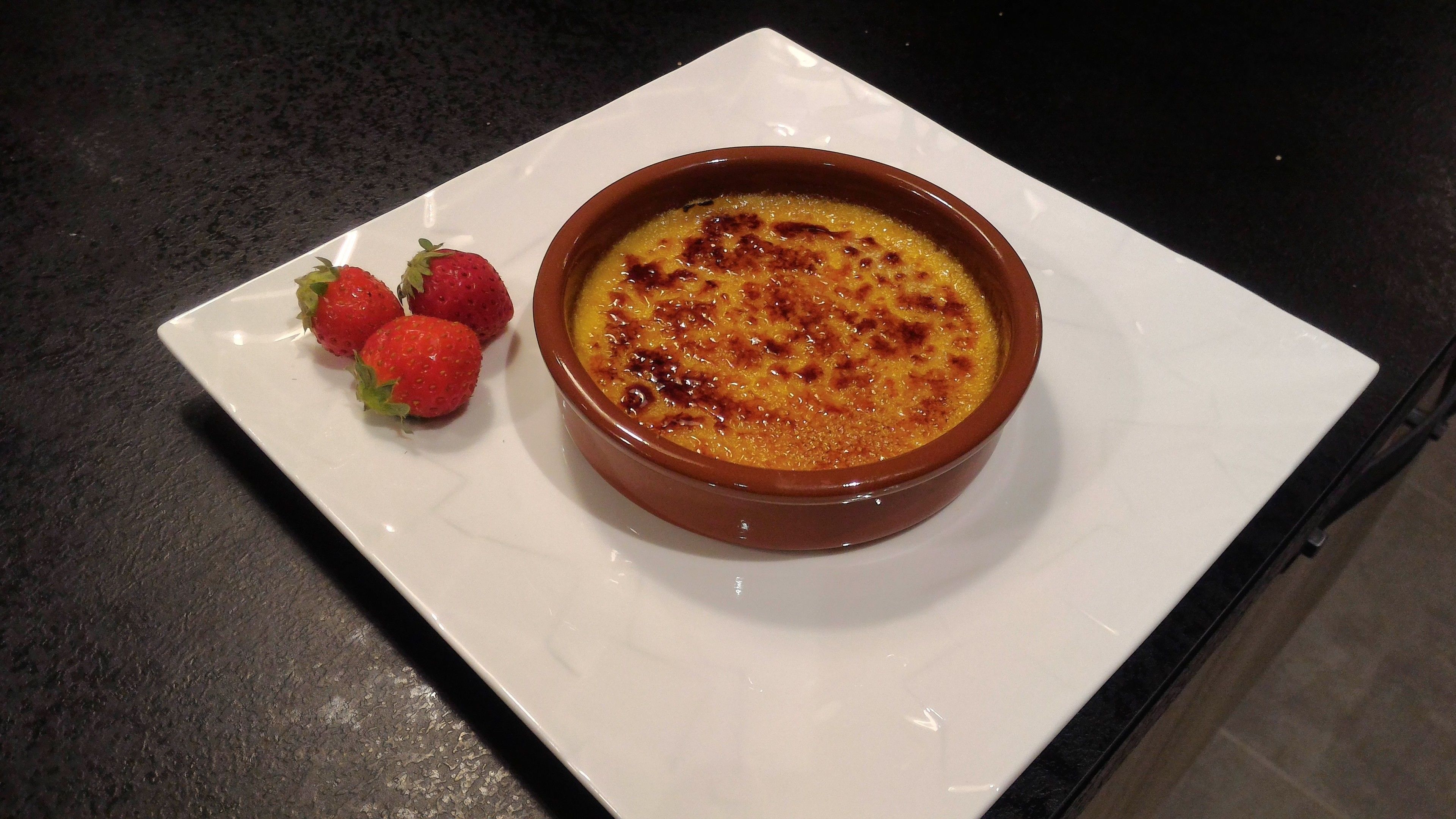Eine Schale Crème brûlée auf einem weißen Teller, daneben drei Erdbeeren.