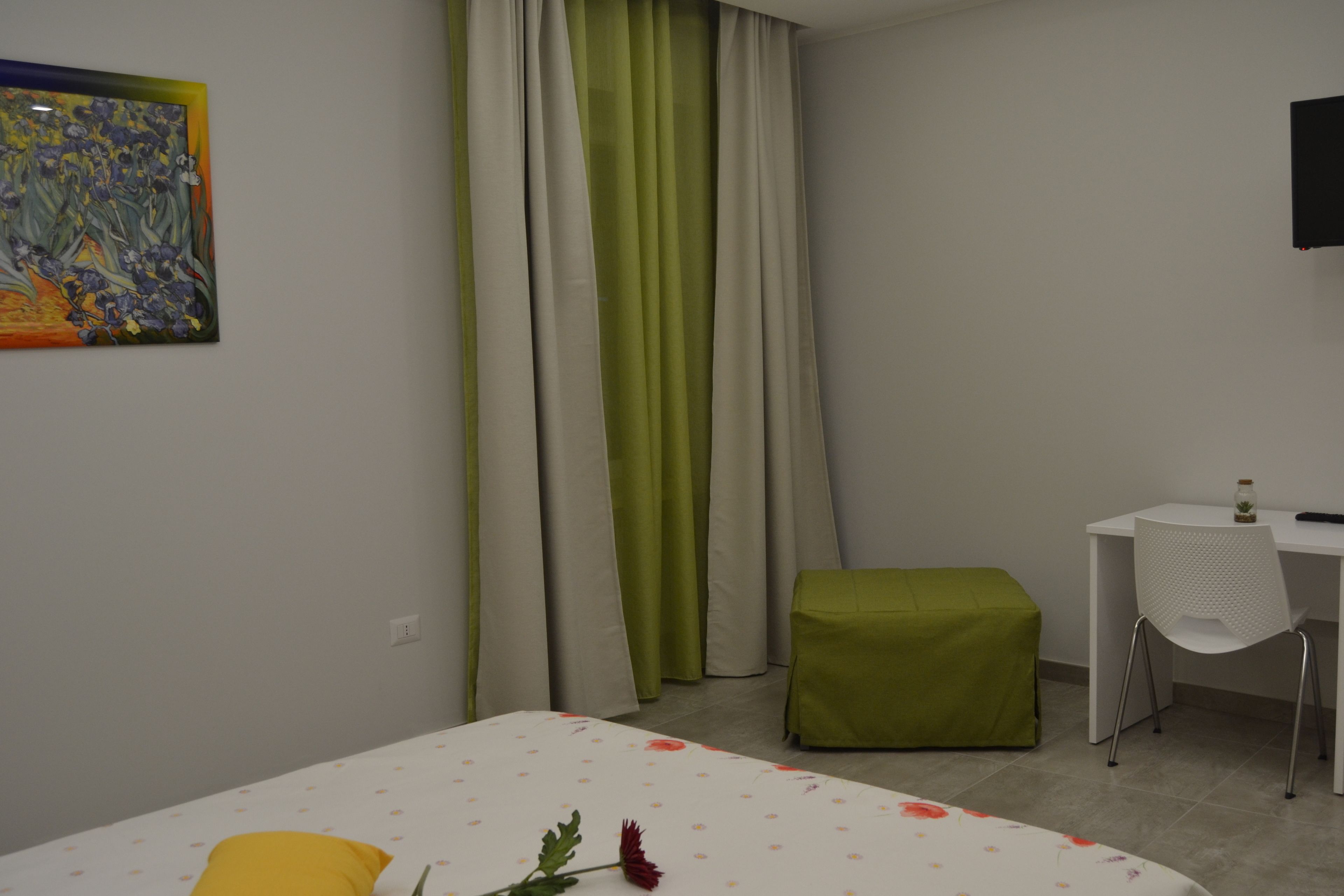 A hotel room with a bed, a desk, a chair, a green ottoman, and a painting on the wall.