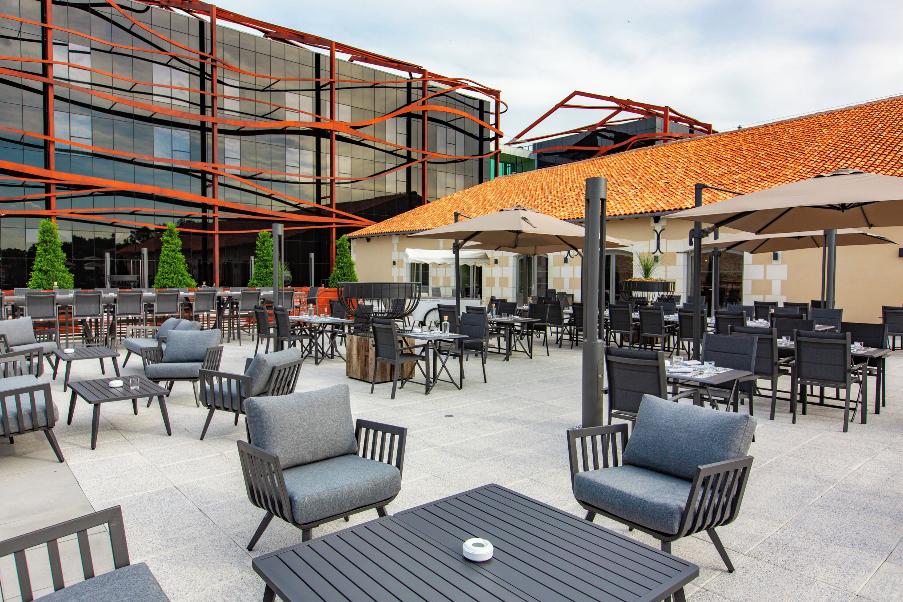An outdoor patio area with multiple tables and chairs, a modern building with glass and metal structures, and a tiled roof.