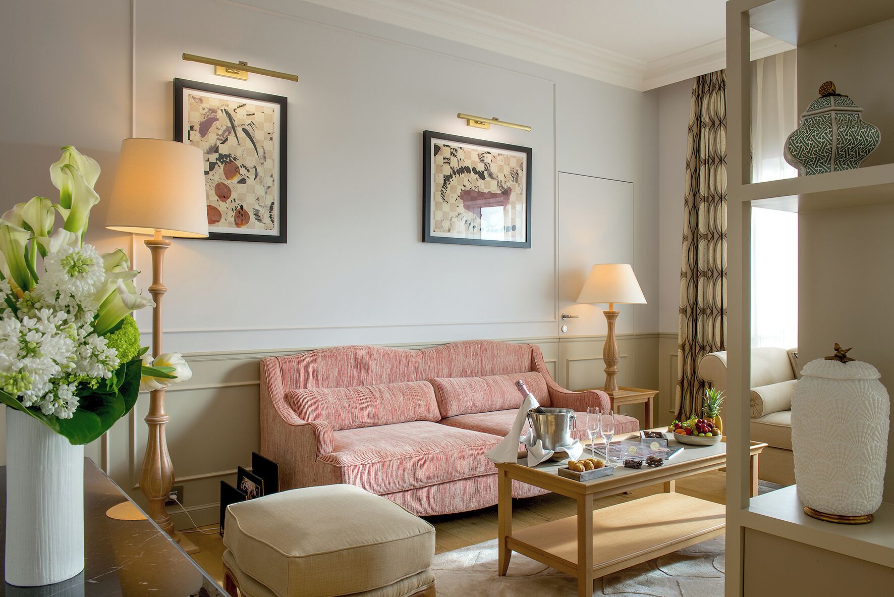 A living room with a pink sofa, a coffee table with a bottle and glasses, a vase with flowers, and two framed artworks on the wall.