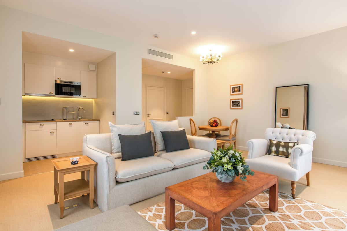 A modern living room with a sofa, armchair, coffee table, and a small side table. There is a kitchen area with cabinets and a microwave. A chandelier hangs from the ceiling, and a mirror is on the wall.