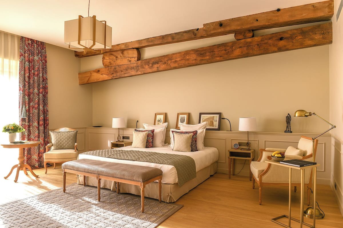 A bedroom with a large bed, a wooden ceiling beam, a bedside table with a lamp, a chair, and a small table with a lamp.