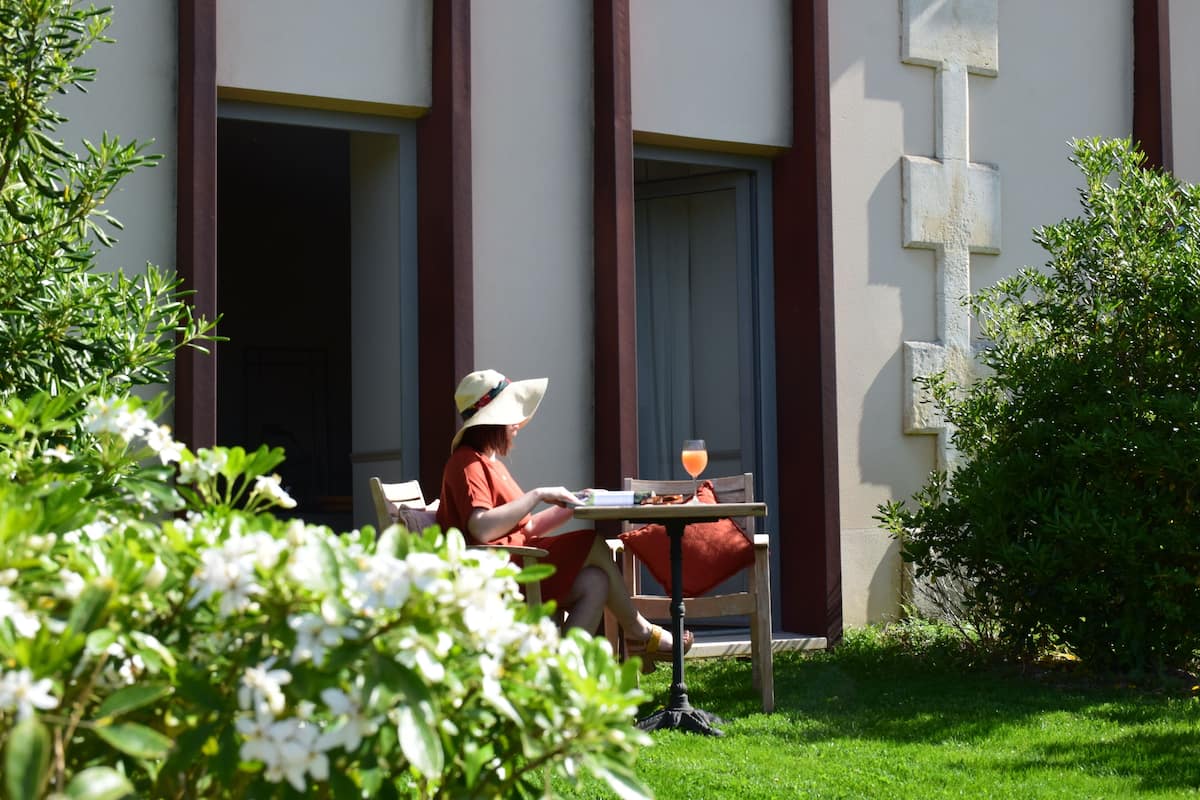 A person sitting at an outdoor table with a drink, surrounded by greenery and a building with large windows and a cross on the wall.