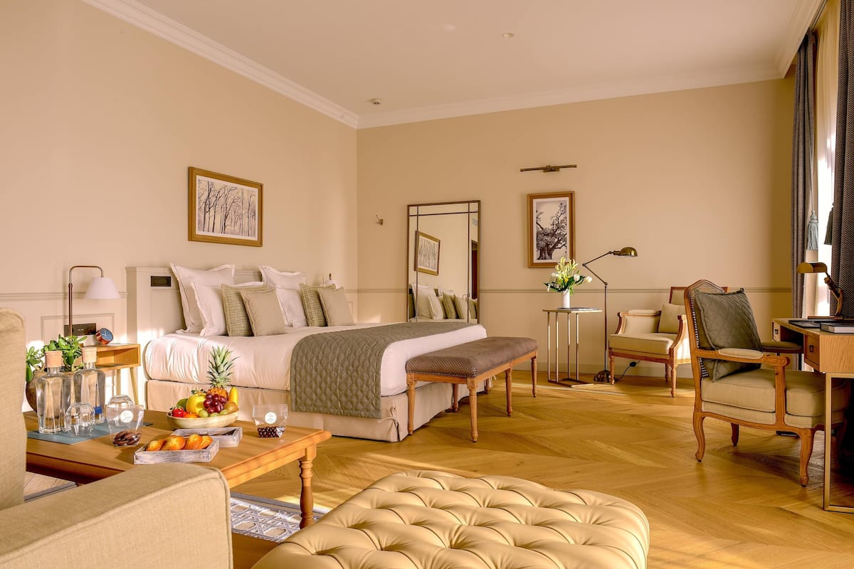 A spacious hotel room with a large bed, a sitting area with a sofa and armchairs, a wooden coffee table, and a small side table with a lamp.