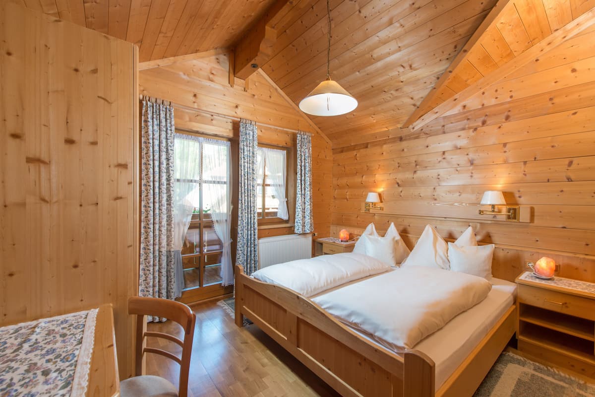 Quarto de cabana de madeira com uma cama de casal, duas mesas de cabeceira com abajures, uma cadeira de madeira e uma janela com cortinas.