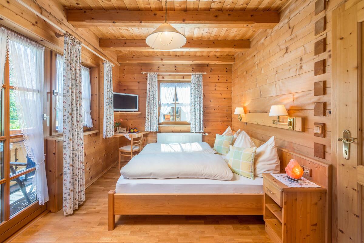Quarto de cabana de madeira com cama, mesa de cabeceira, abajur e televisão na parede.
