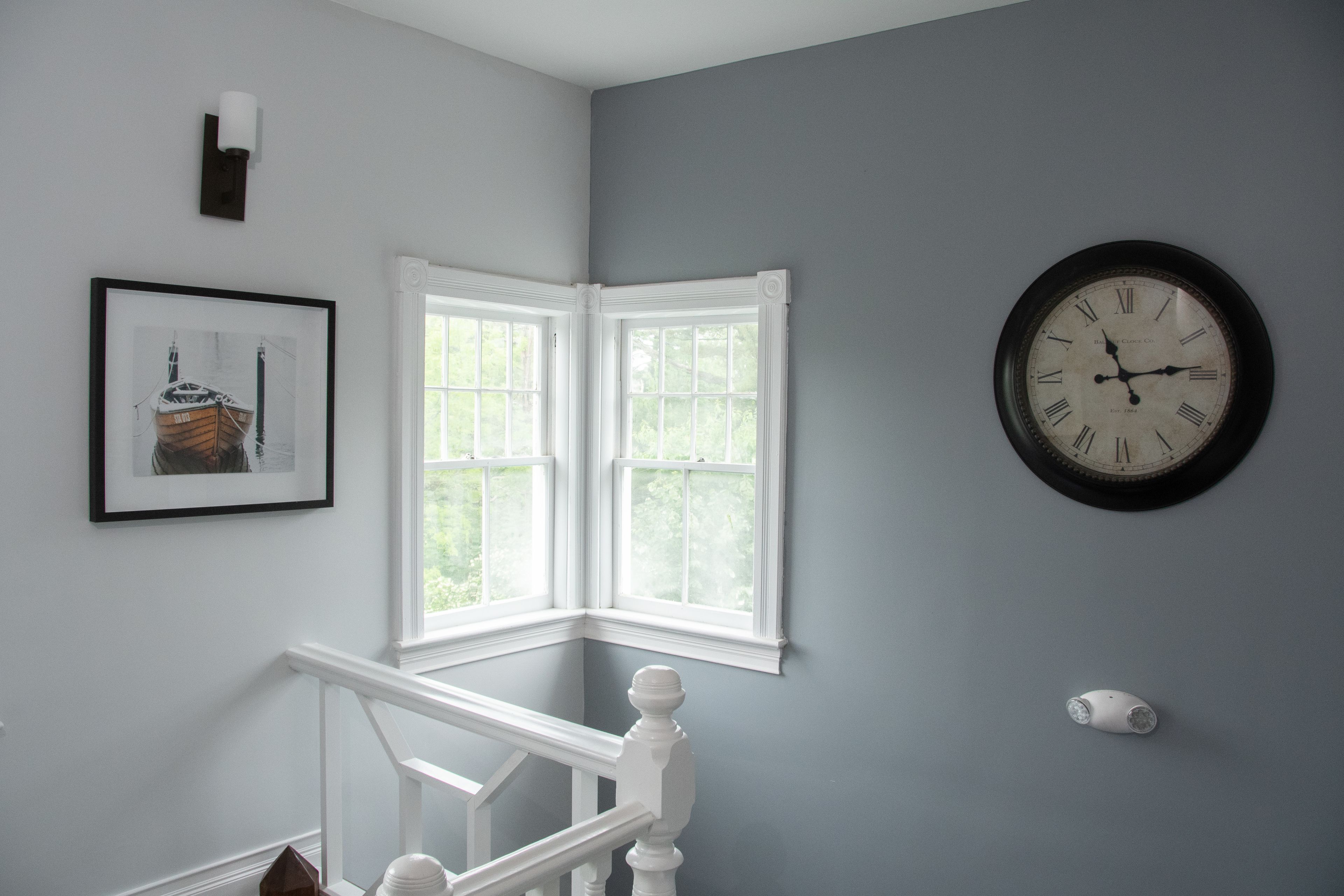 A corner room with a white staircase, a framed picture, a wall clock, and a window with white trim.