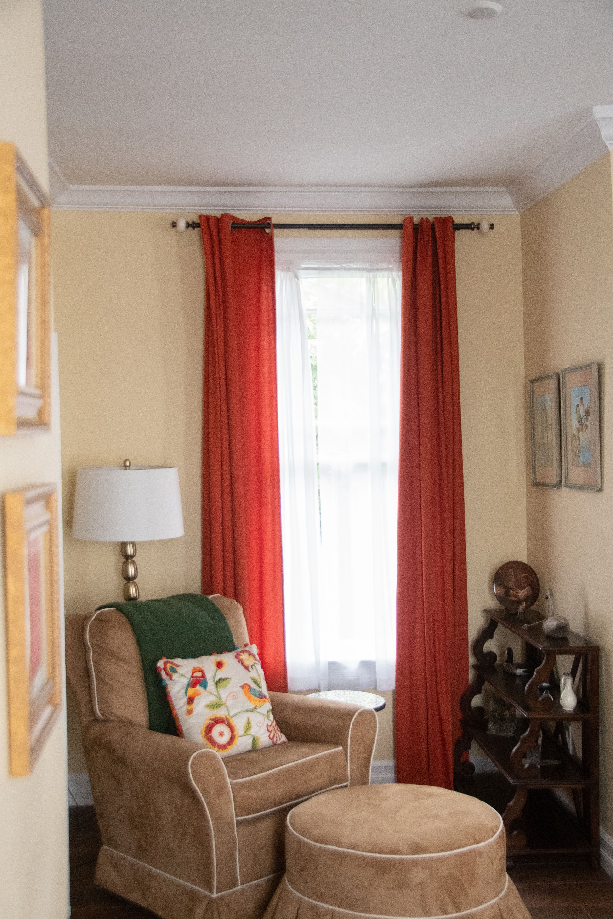 A room with a brown armchair, a small ottoman, a lamp, and a window with red curtains.