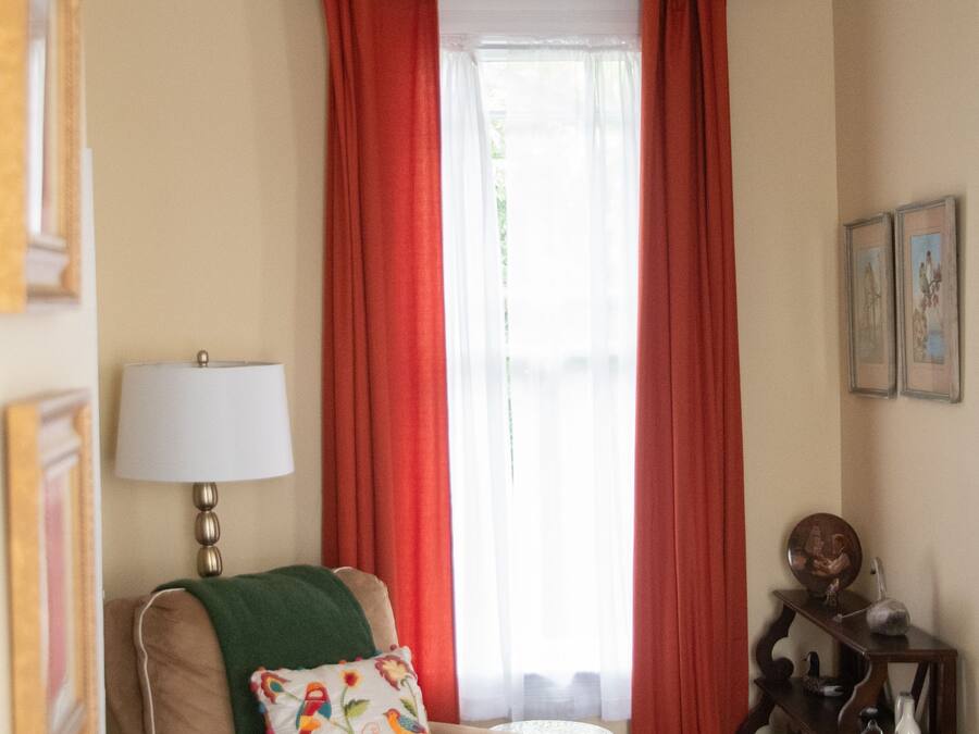A room with a brown armchair, a small ottoman, a lamp, and a window with red curtains.