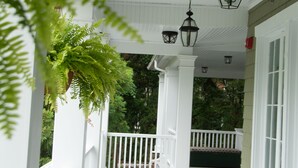 Un porche cubierto con barandillas y columnas blancas, faroles colgantes y una planta verde.