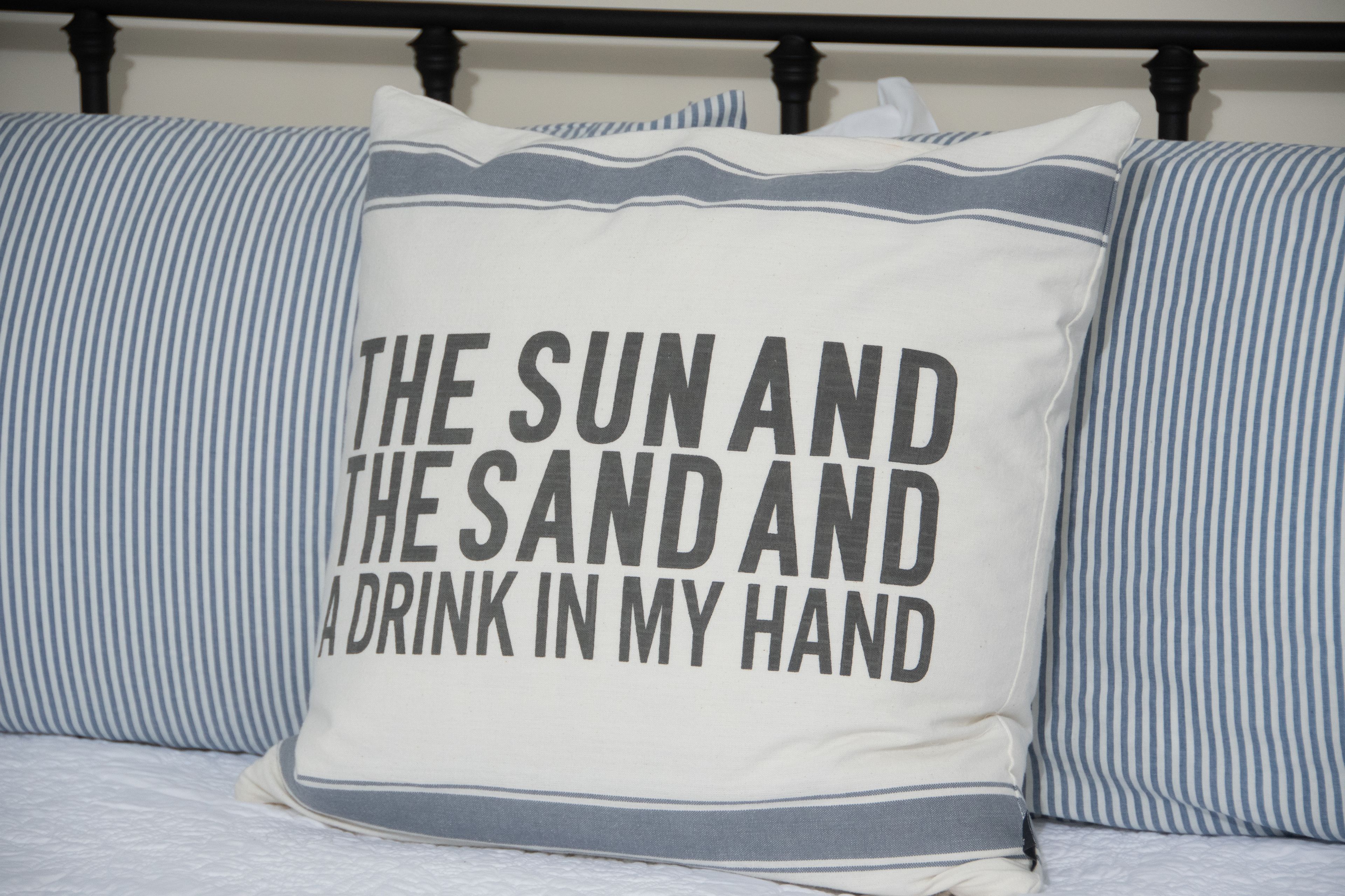 A pillow with the text "THE SUN AND THE SAND AND A DRINK IN MY HAND" on a bed with striped bedding.