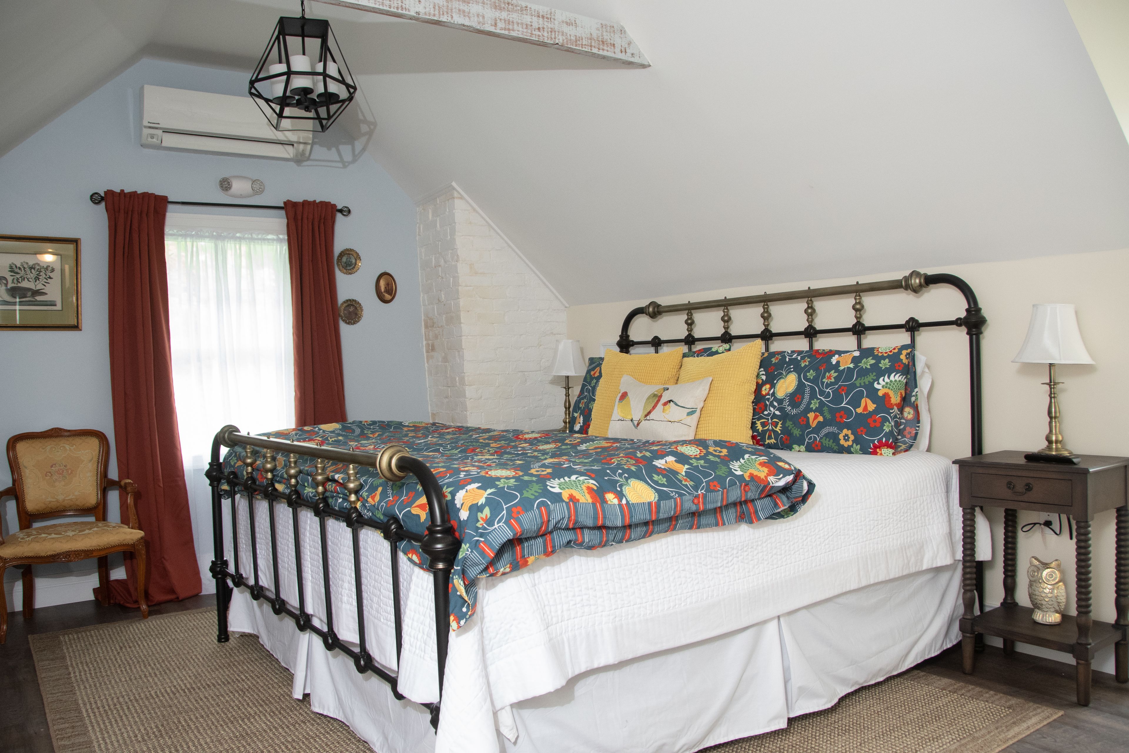 A bedroom with a bed, a chair, a nightstand, a lamp, and a wall-mounted air conditioner.